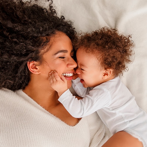 A woman and her baby smile joyfully while lying together on a cozy bed, capturing a moment of warmth and affection